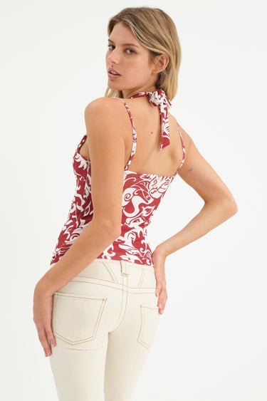 Woman wearing a demi cup halter top with bra strap details in maroon-and-white hawaiian print with off white jeans on a white background